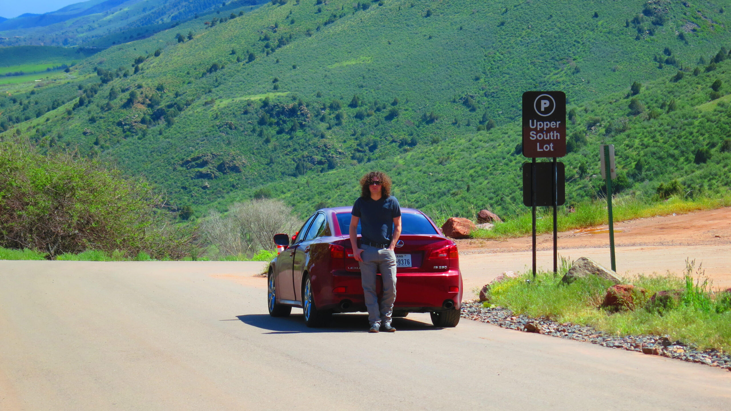 A person stands on the side of a road with a mountainous landscape in the background, next to a red car parked by a sign indicating directions to various locations.