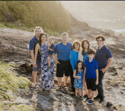 The image shows a family posing for a photo on a rocky shoreline with a scenic background.