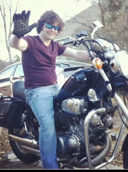 A man wearing sunglasses is sitting on a parked motorcycle, waving at the camera with one hand.