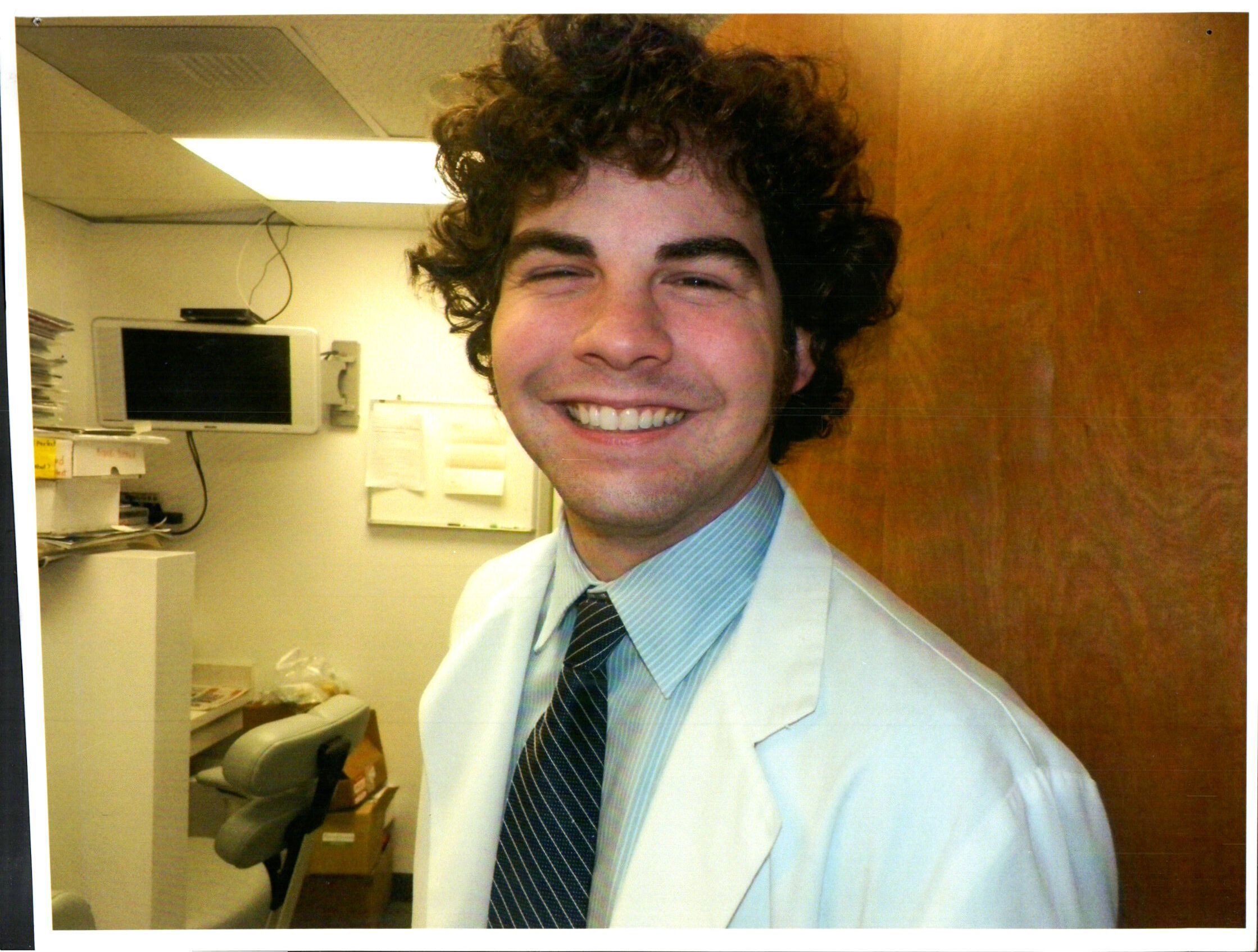 The image shows a young man with curly hair wearing a white lab coat, smiling at the camera, standing indoors with medical equipment and a desk in the background.