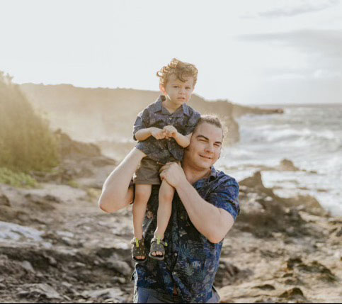 The image depicts a man holding a child on a beach with ocean waves in the background.