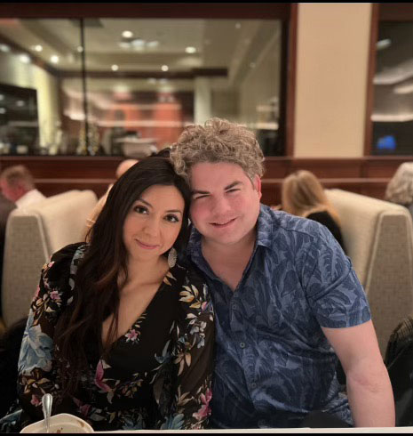 The image shows a man and a woman sitting closely together at a dining table, both smiling and looking towards the camera.