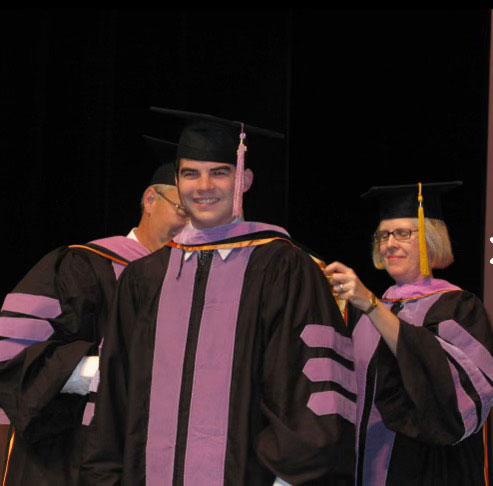 The image features a graduation ceremony where a male graduate in academic regalia is receiving an award from two individuals wearing similar attire, with one adjusting his medal.