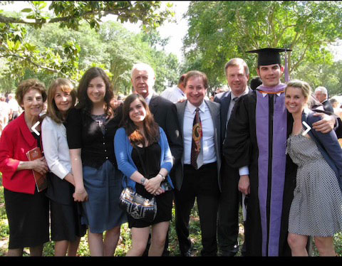 The image shows a group of people posing together outdoors  they are dressed formally with some wearing academic regalia, suggesting an event such as a graduation ceremony.