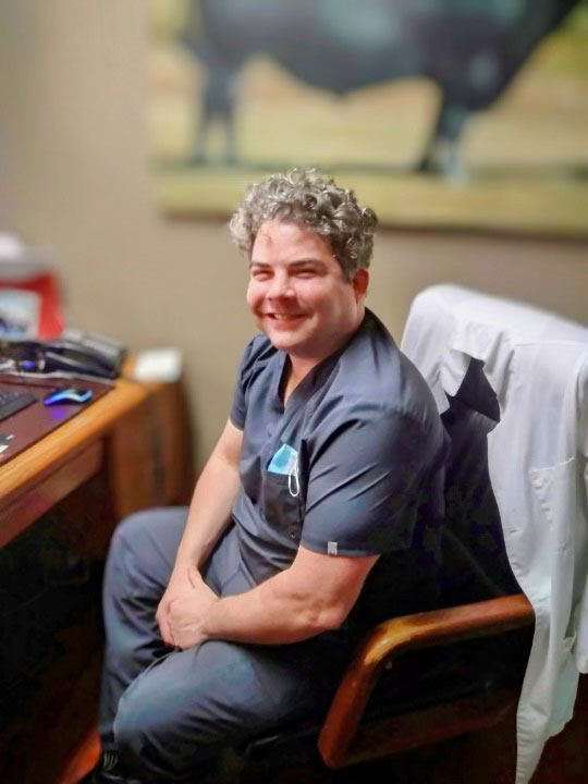 The image depicts a man sitting at a desk with a laptop, smiling towards the camera.