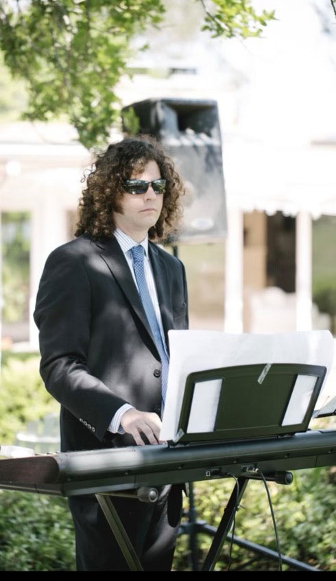 The image shows a man standing behind a keyboard at an outdoor event, wearing sunglasses and a suit, playing music on a stage with trees in the background.