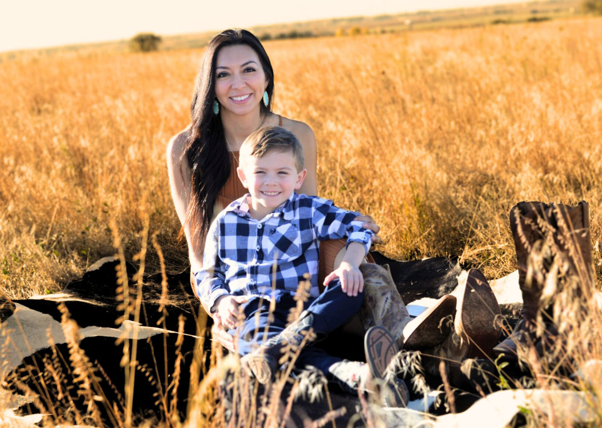 A woman and a young boy are sitting together on a pile of construction materials in an outdoor setting with tall grasses.