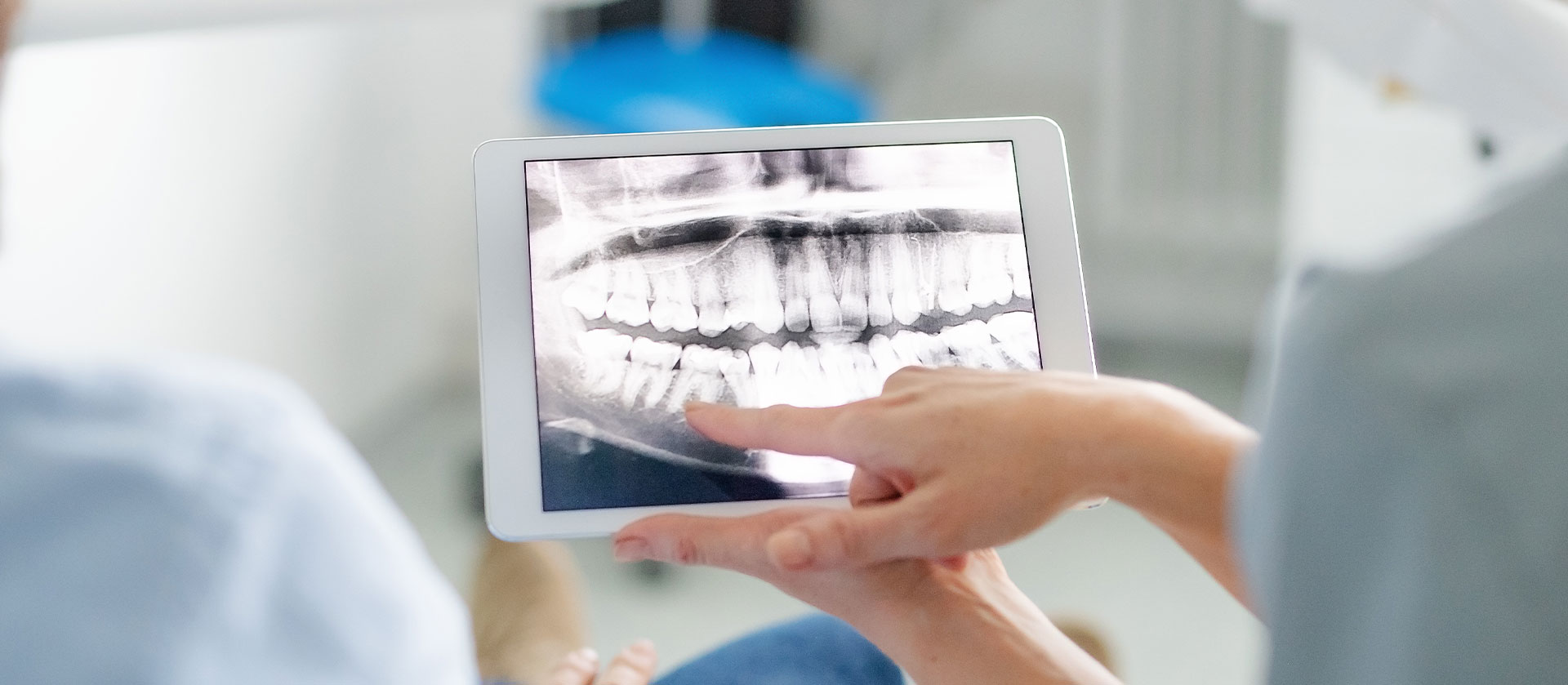 A person holding a tablet with an X-ray displayed on the screen while another individual appears to be working in a dental or medical setting, both are wearing scrubs.