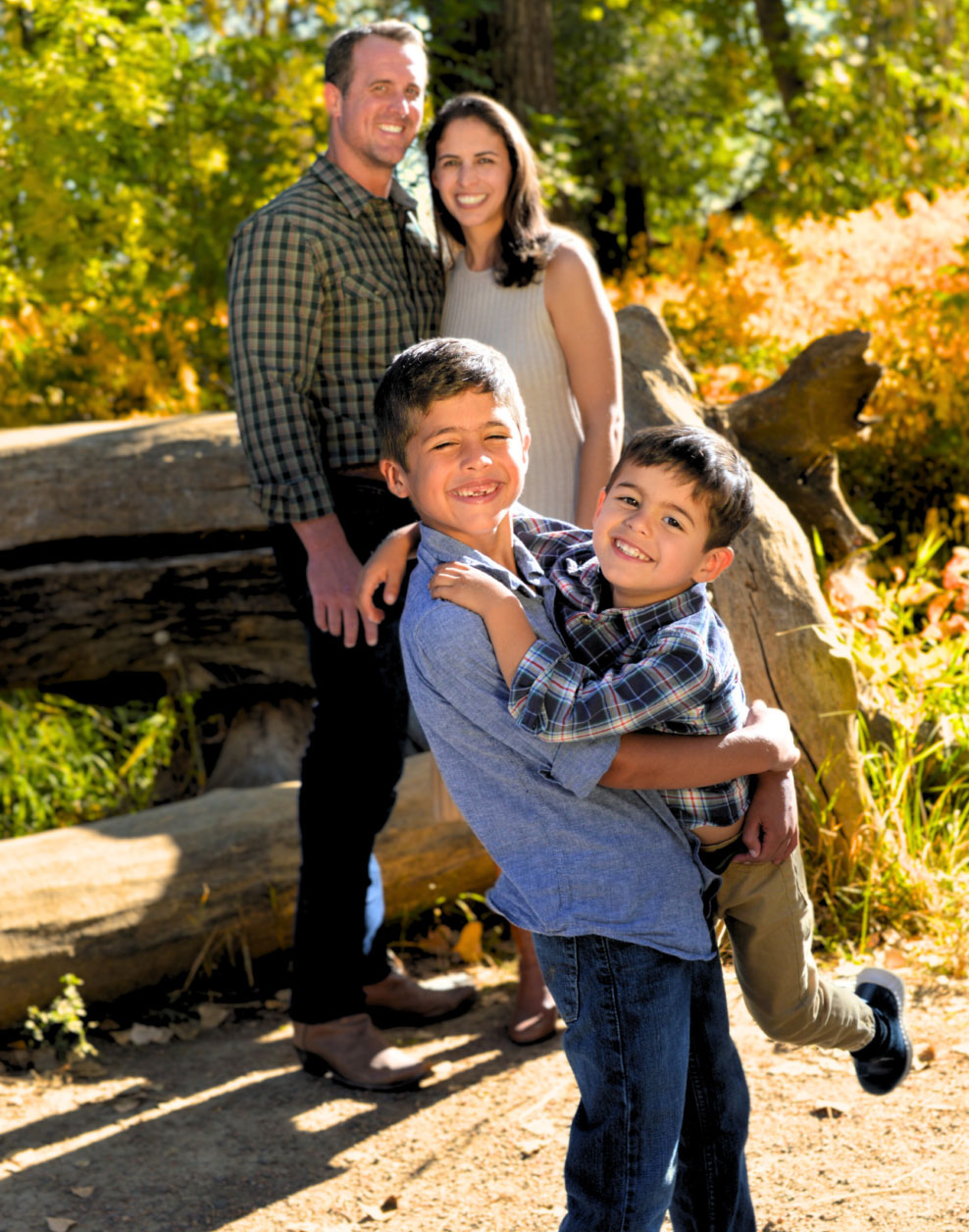 A family poses for a photo outdoors during autumn, with a man holding two young boys while a woman stands behind them.